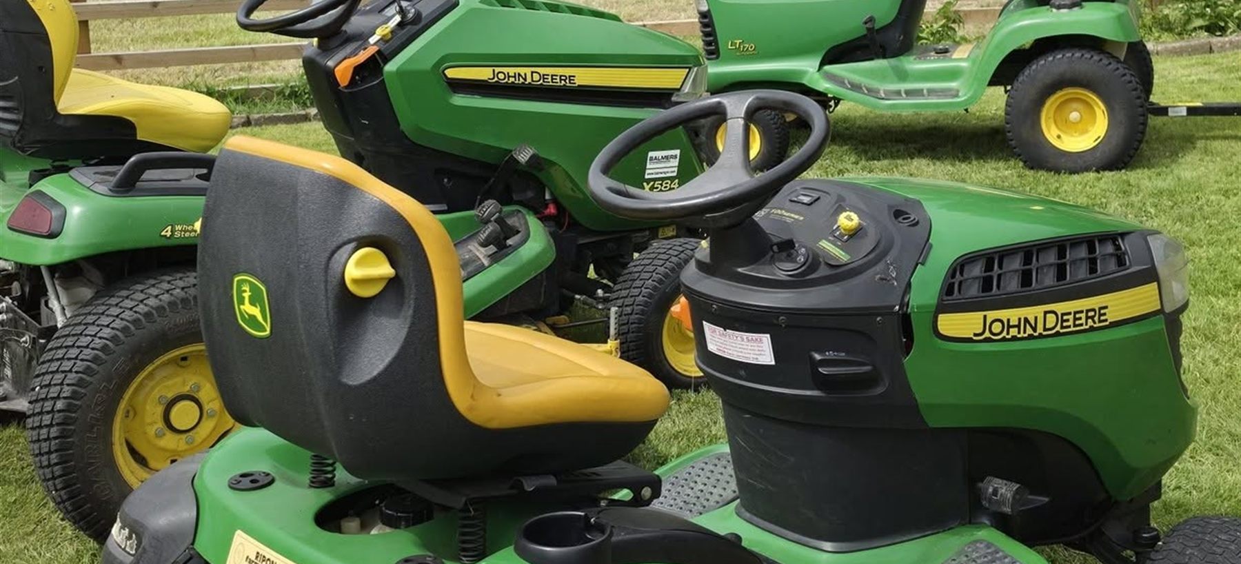 Three John Deere ride-on mowers lined up on grass, in for servicing